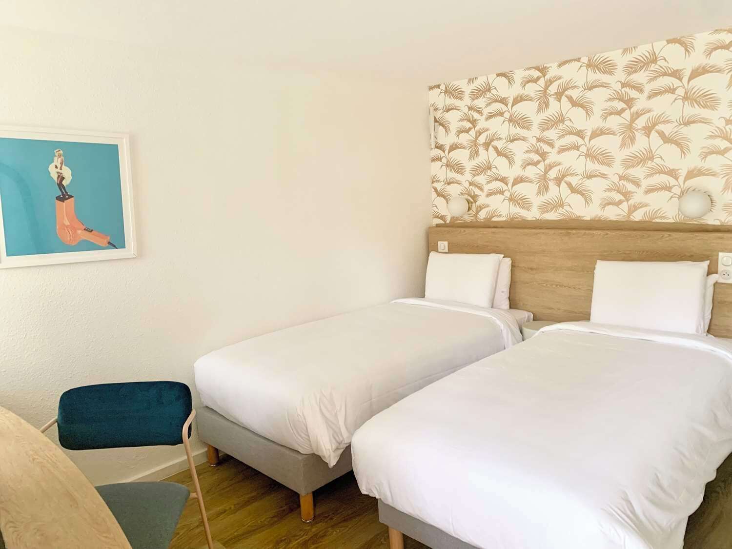 A hotel room with two beds, a wooden headboard, a wall with a palm leaf pattern, and a framed picture on the wall.