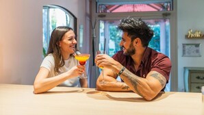Dos personas sentadas en una mesa con bebidas, una ventana y una puerta al fondo.