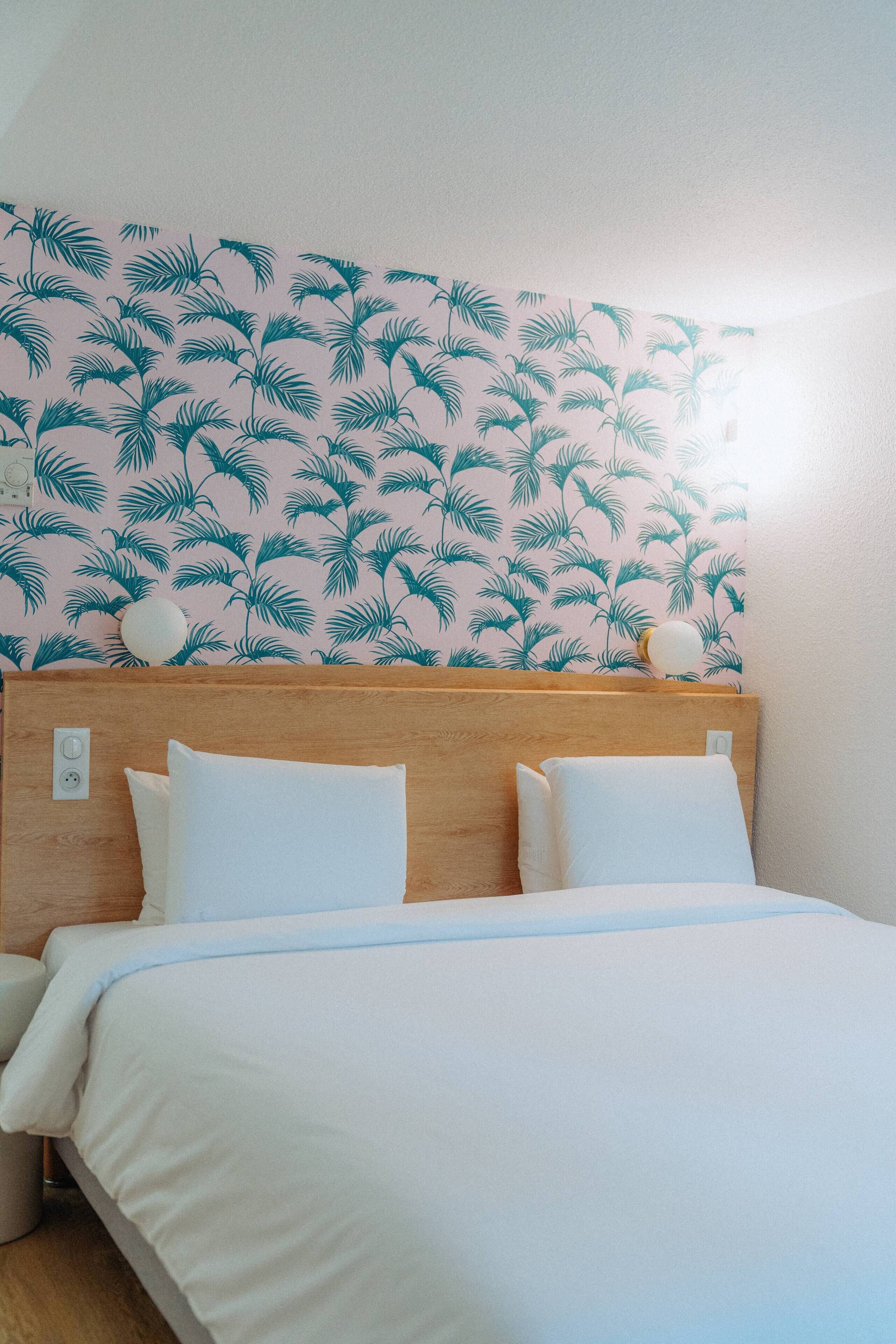 Una cama con ropa blanca, un cabecero de madera y una pared con un diseño de hojas tropicales.