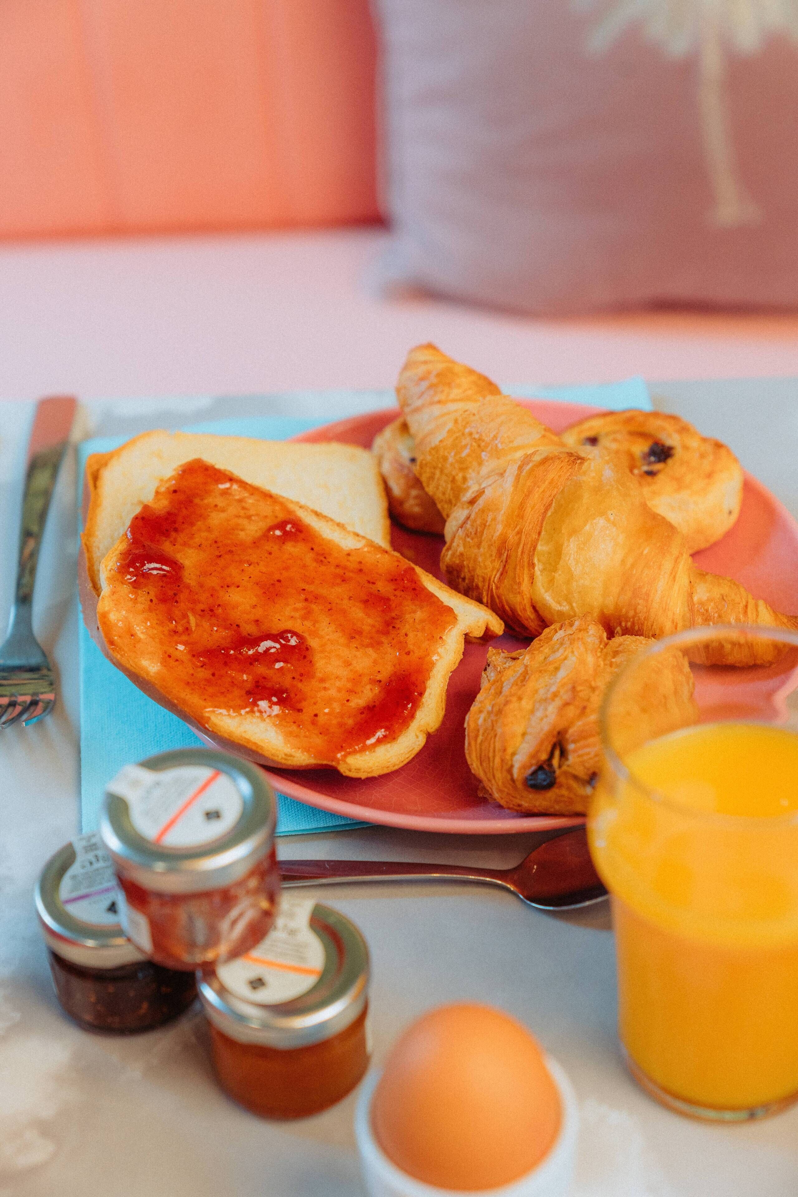 A breakfast plate with croissants, toast with jam, a glass of orange juice, a boiled egg, and condiments.