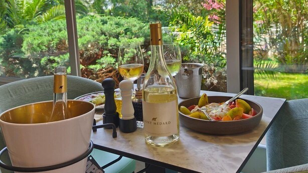 Una mesa con una botella de vino Saint Médard, copas, un bol de fruta y un molinillo de pimienta.
