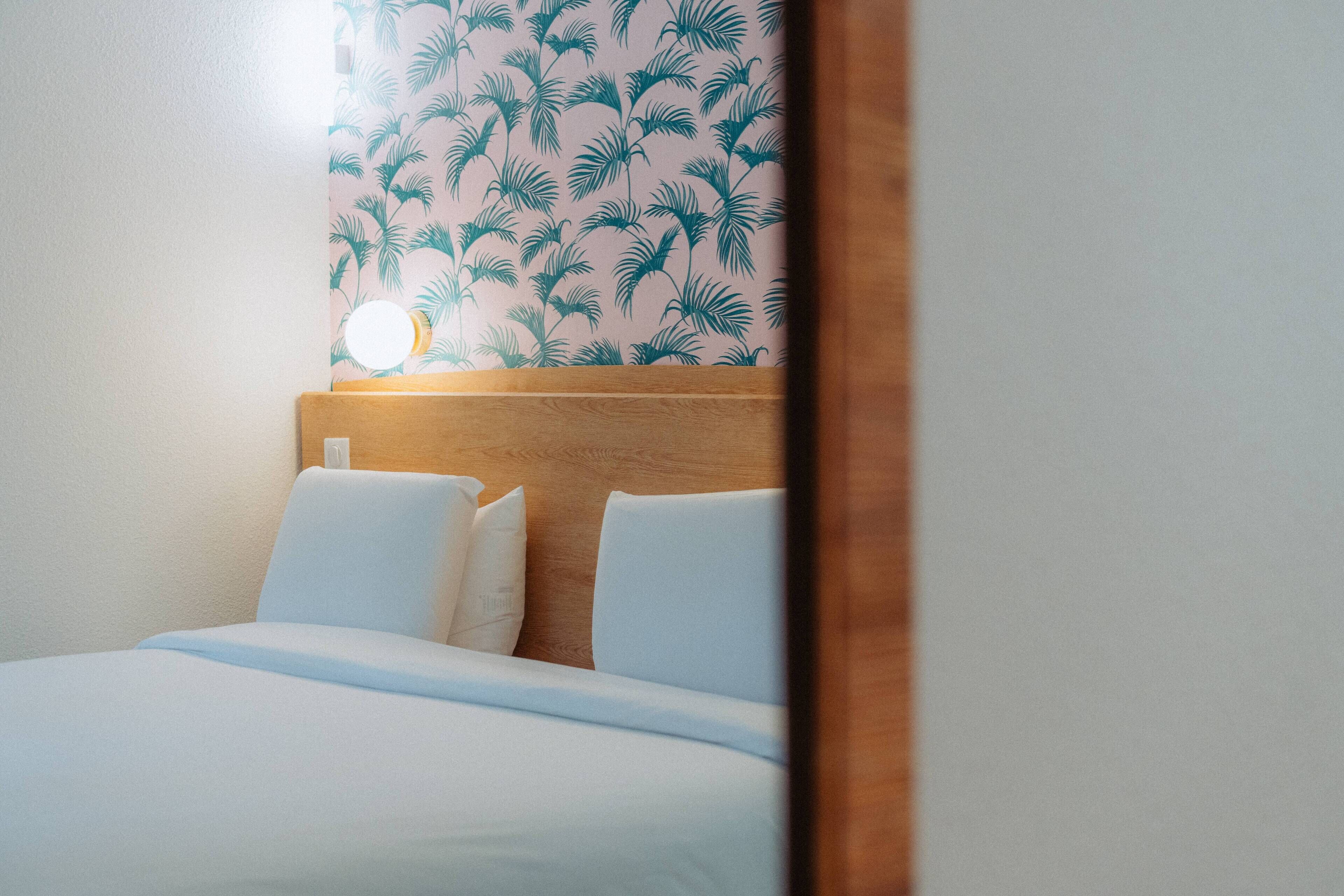A hotel room with a bed, white bedding, a wooden headboard, and a wall with a tropical leaf pattern.