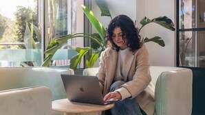 Una persona sentada en un sofá usando un ordenador portátil en una habitación con ventanas amplias y plantas en macetas.
