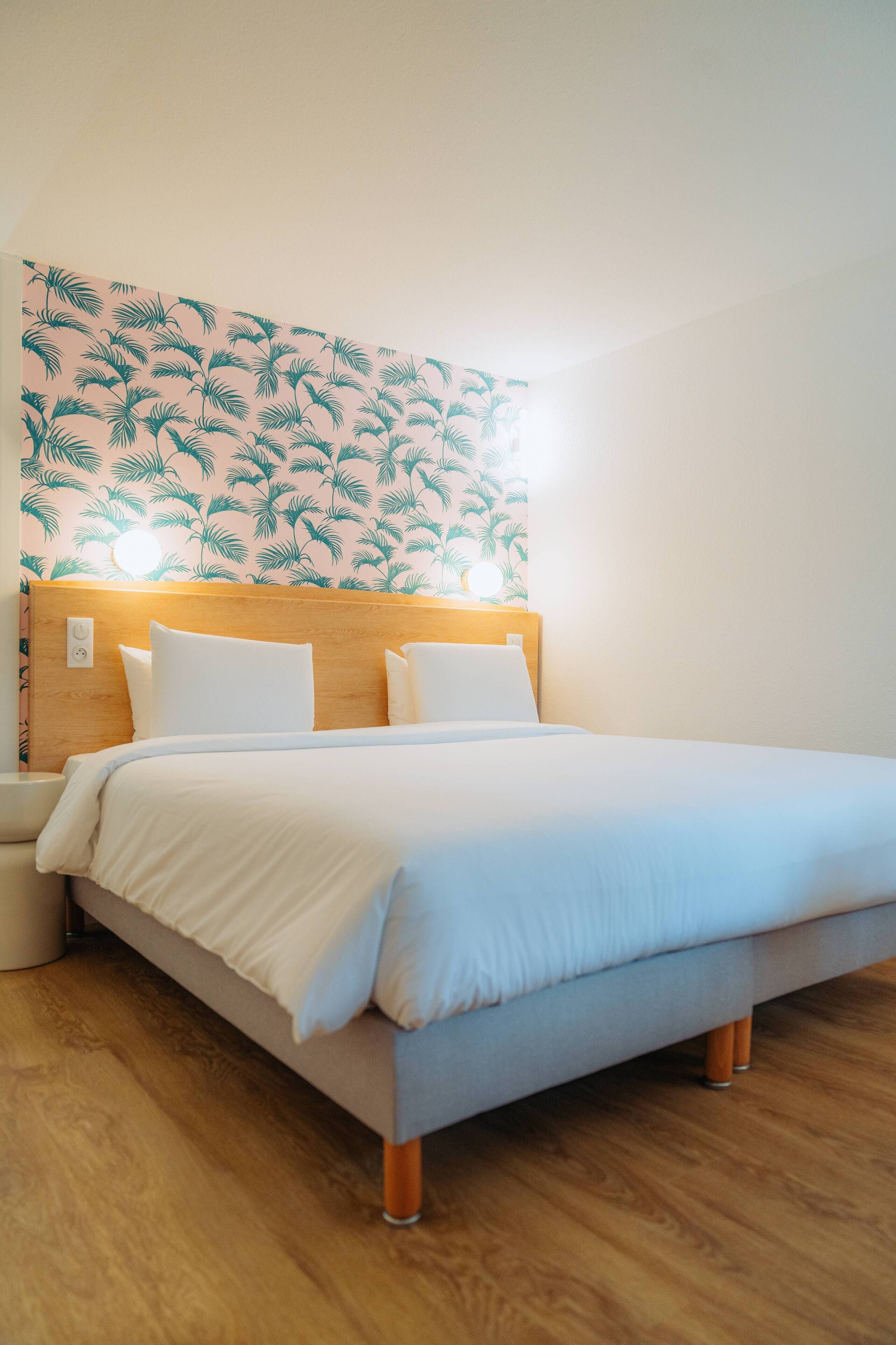 Ein ordentlich bezogenes Bett mit Holz Kopfteil, weißer Bettwäsche und einer Wand mit tropischem Blattmuster.