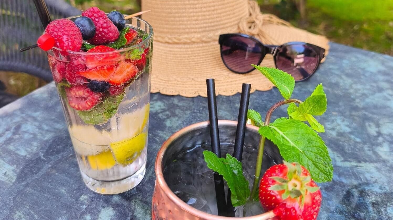 A glass with fruit and a straw, a copper mug with a straw, sunglasses, and a straw hat on a table.