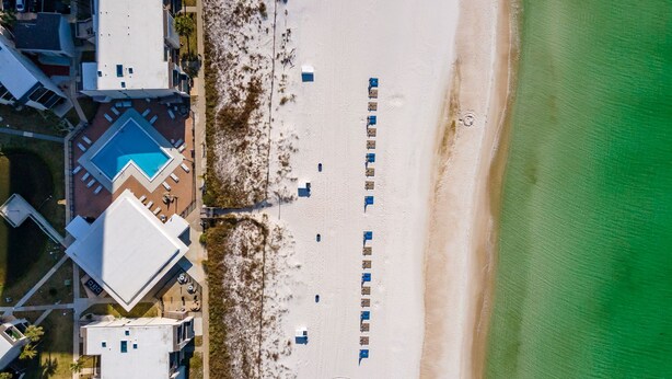 An aerial view of a beach with a row of blue beach umbrellas, a swimming pool, and adjacent buildings. - Family-friendly oceanfront escape w/ views steps from private beach (Panama City Beach)