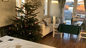 Une pièce chaleureuse, décorée d’un sapin de Noël, avec une table à manger recouverte d’une nappe verte et un lit agrémenté d’une couvre-lit blanche.