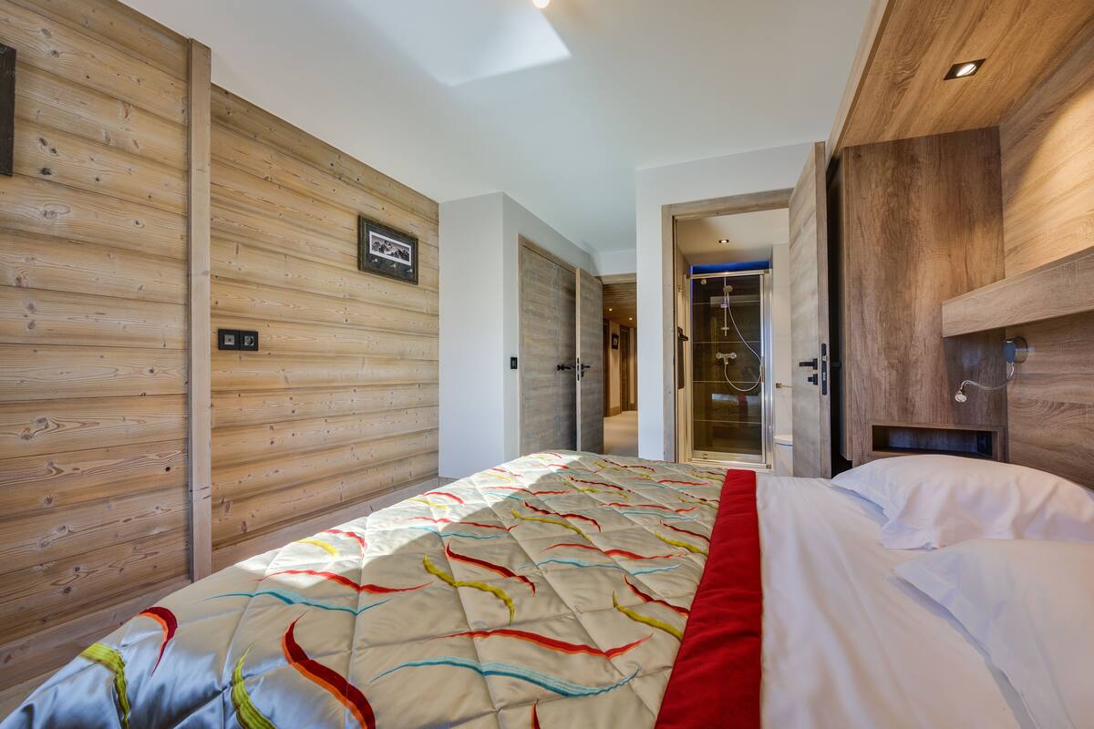 Moderni hotellihuone, jossa on puinen seinä, värikäs peitto sängyllä ja avoimesta ovesta näkyvä suihkutila.