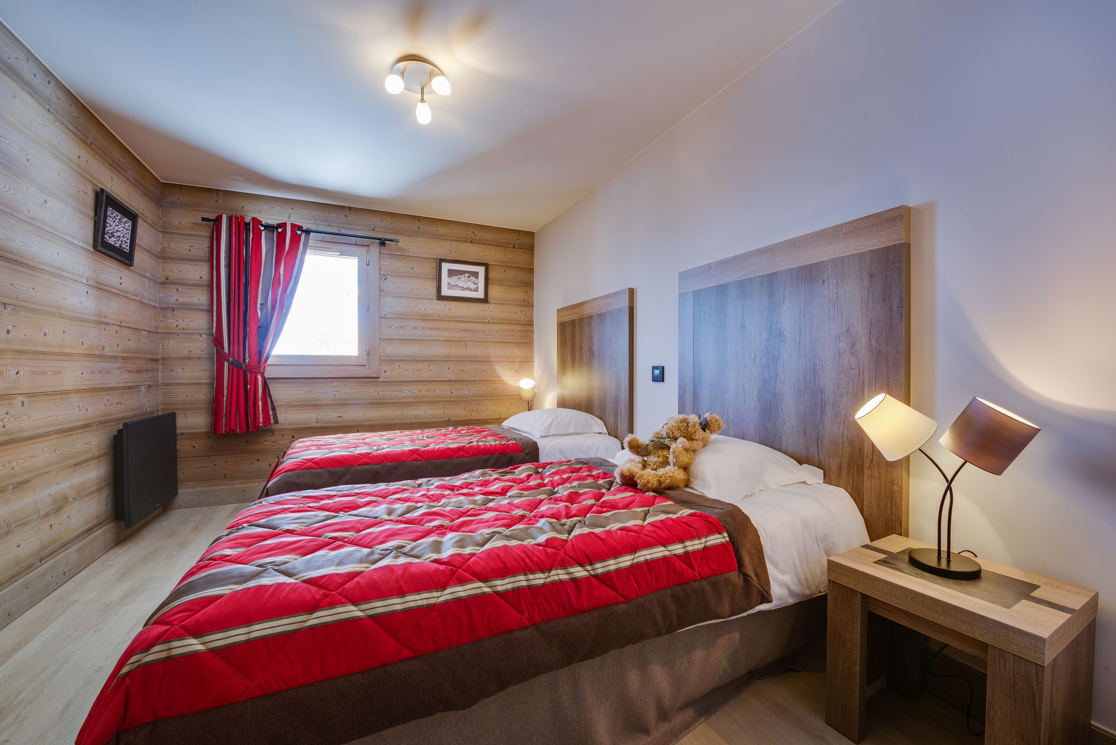 A wooden cabin room with two beds, a window with red curtains, a teddy bear, and a nightstand with a lamp.