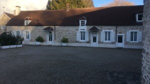 Ein Gebäude aus Stein mit mehreren Fenstern und Türen, einem Kieshof und einem großen Baum im Hintergrund.