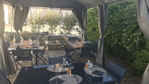 Eine Terrasse im Freien mit Tischen und Stühlen, einige davon mit Getränken und Gläsern, unter einem überdachten Bereich mit Vorhängen.