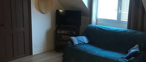 A room with a sloped ceiling, a dark wooden door, a television on a wooden stand, a blue sofa, and a window with blinds.
