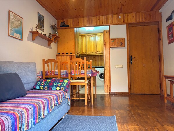 Un salon chaleureux avec un canapé, des fauteuils, un tapis, un plancher en bois, une table à manger et un coin cuisine doté de meubles en bois et d’un lave-linge.