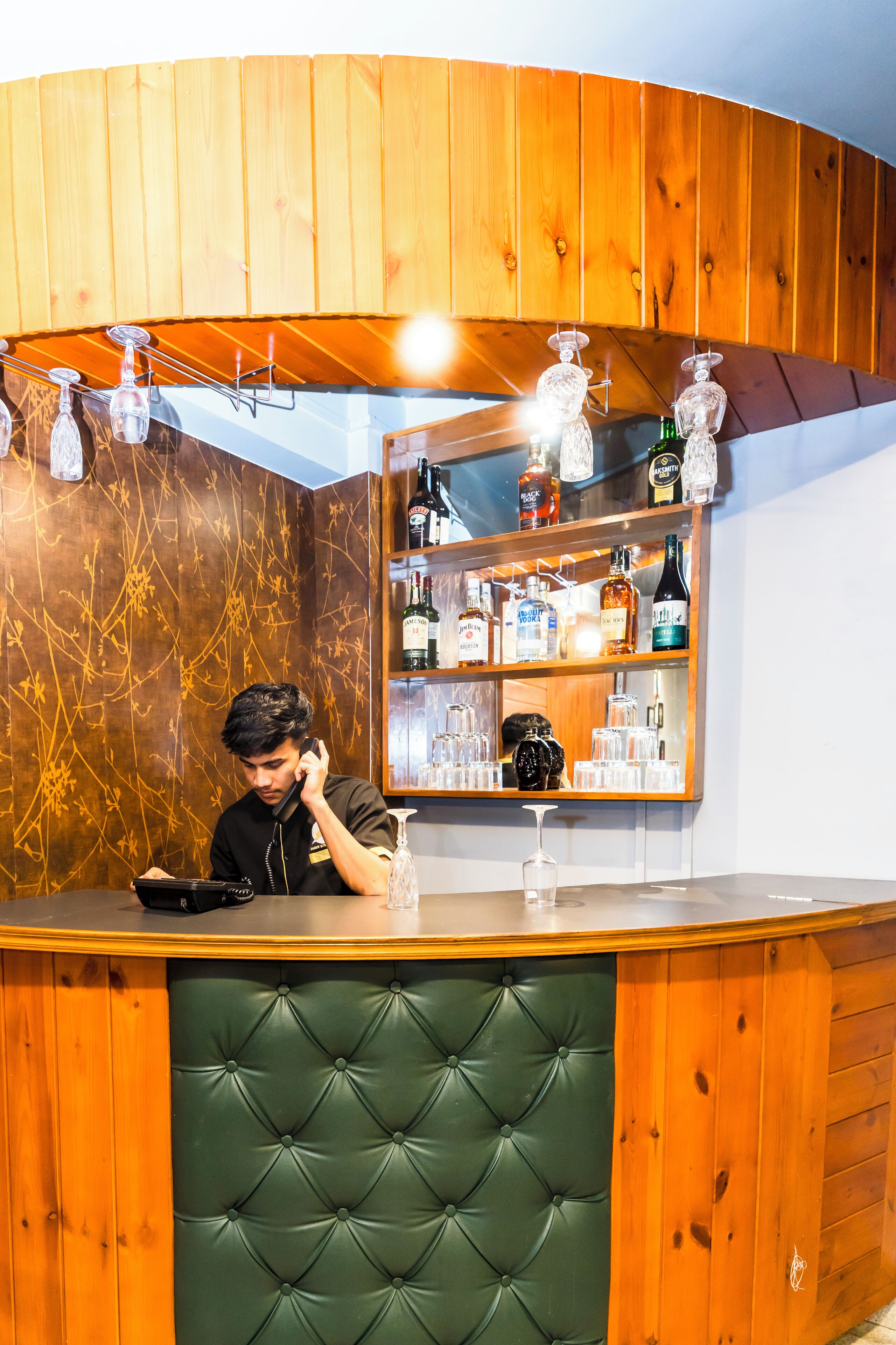 A bar area with wooden paneling, a green cushioned bar counter, and a mirrored wall displaying various bottles and glasses.