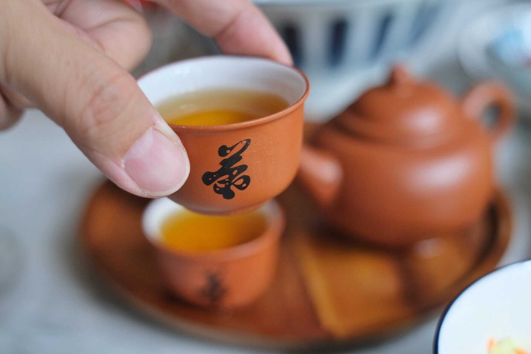Una mano sosteniendo una taza con diseño, una tetera y un platito sobre una mesa.