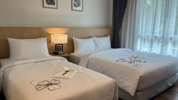 A hotel room with two beds, a wooden headboard, a nightstand with a lamp, and a window with sheer curtains.