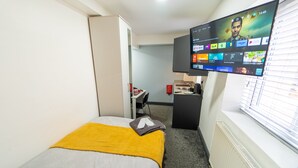 Quarto de hotel com cama, televisão fixada na parede exibindo diversos canais, escrivaninha com cadeira e janela com persianas.