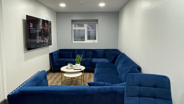 Quarto com sofá modular azul, uma mesinha redonda com uma planta e uma TV fixada na parede.