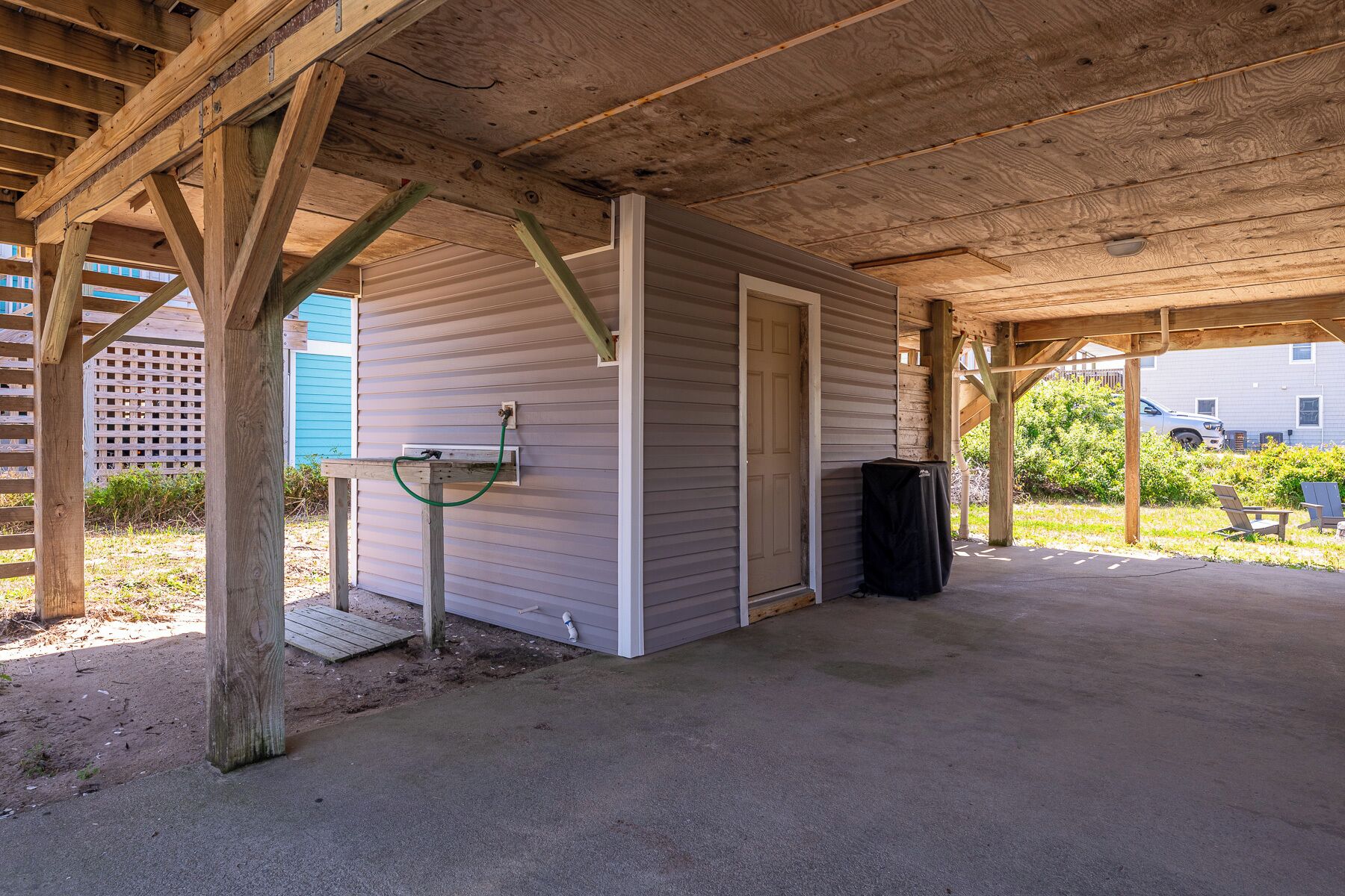 A covered outdoor area with a concrete floor, a wooden structure with a door, and a trash bin.