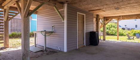 A covered outdoor area with a concrete floor, a wooden structure with a door, and a trash bin.