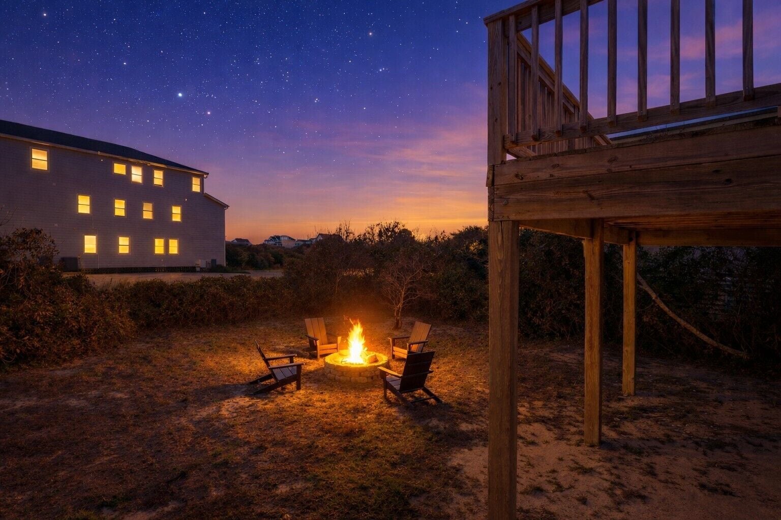 A wooden deck with a fire pit and chairs, a white building with multiple windows, and a starry night sky.