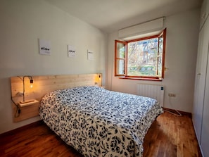 Une chambre avec une tête de lit en bois, un lit recouvert d’une couvre-lit à motifs floraux, une lampe de chevet, une fenêtre donnant sur des immeubles et un radiateur.