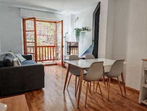 Une pièce avec un plancher en bois, un canapé, une table à manger avec des chaises, une plante sur une étagère et une vue à travers une porte vitrée.
