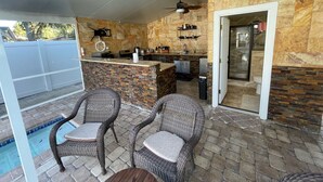 Un área de cocina al aire libre con paredes de piedra, mobiliario de mimbre y una piscina.