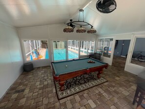 Ein Poolraum mit Billardtisch, Deckenventilator und Blick auf ein Außenbecken.