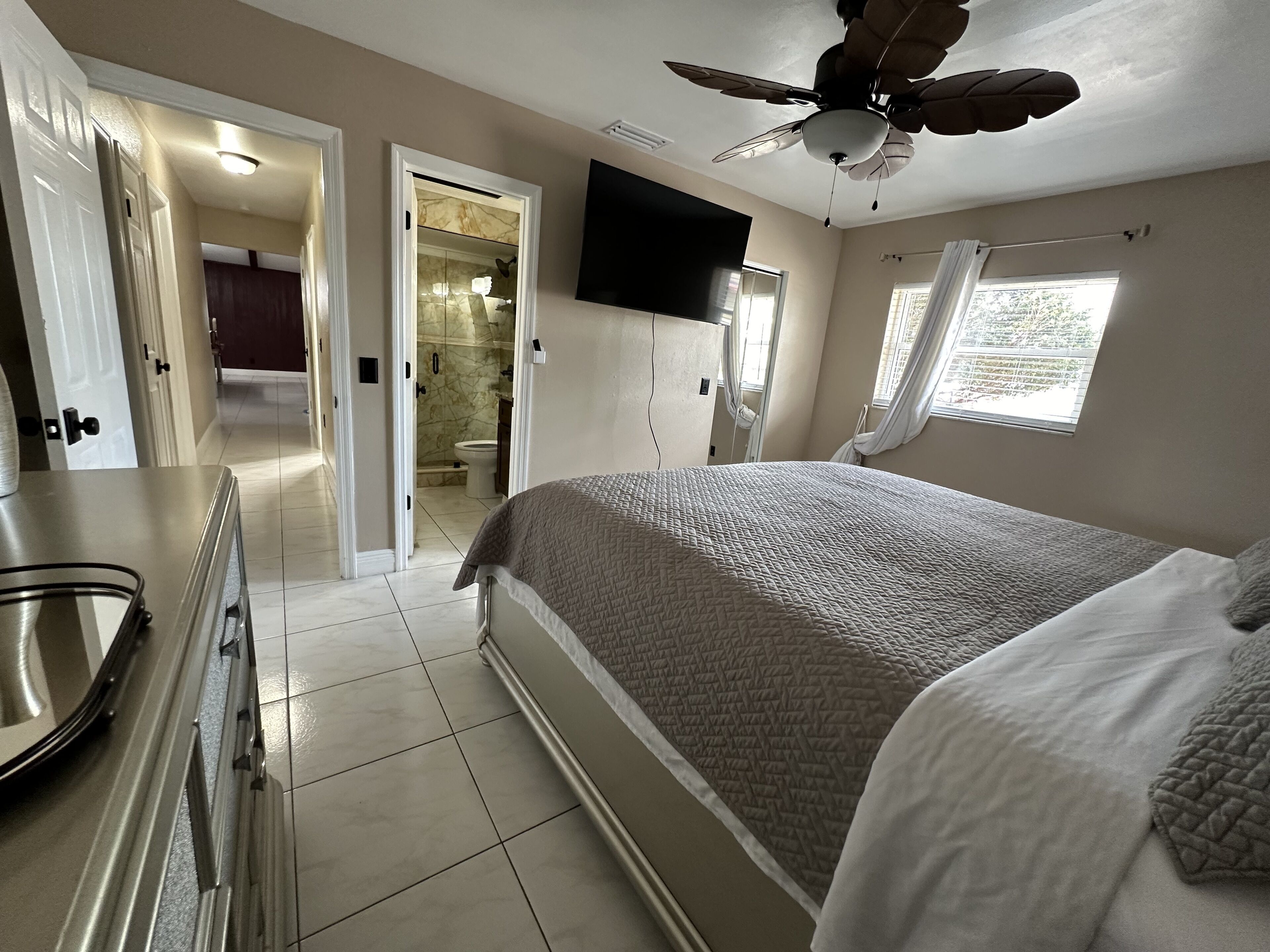 A bedroom with a bed, a ceiling fan, a TV mounted on the wall, and a window with curtains.