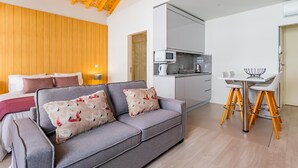Una sala moderna con un sofá gris, vigas de techo de madera, una cocina con armarios blancos y un comedor con mesa blanca y sillas amarillas.