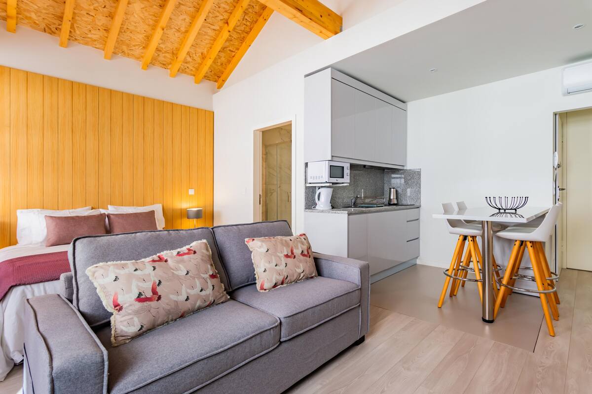 A modern living room with a grey sofa, wooden ceiling beams, a kitchen with white cabinets, and a dining area with a white table and yellow chairs.