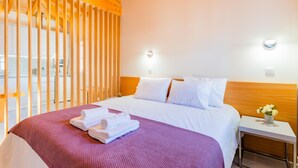 Habitación de hotel con techo de madera, cama con ropa blanca, manta morada y mesita de noche con un florero.