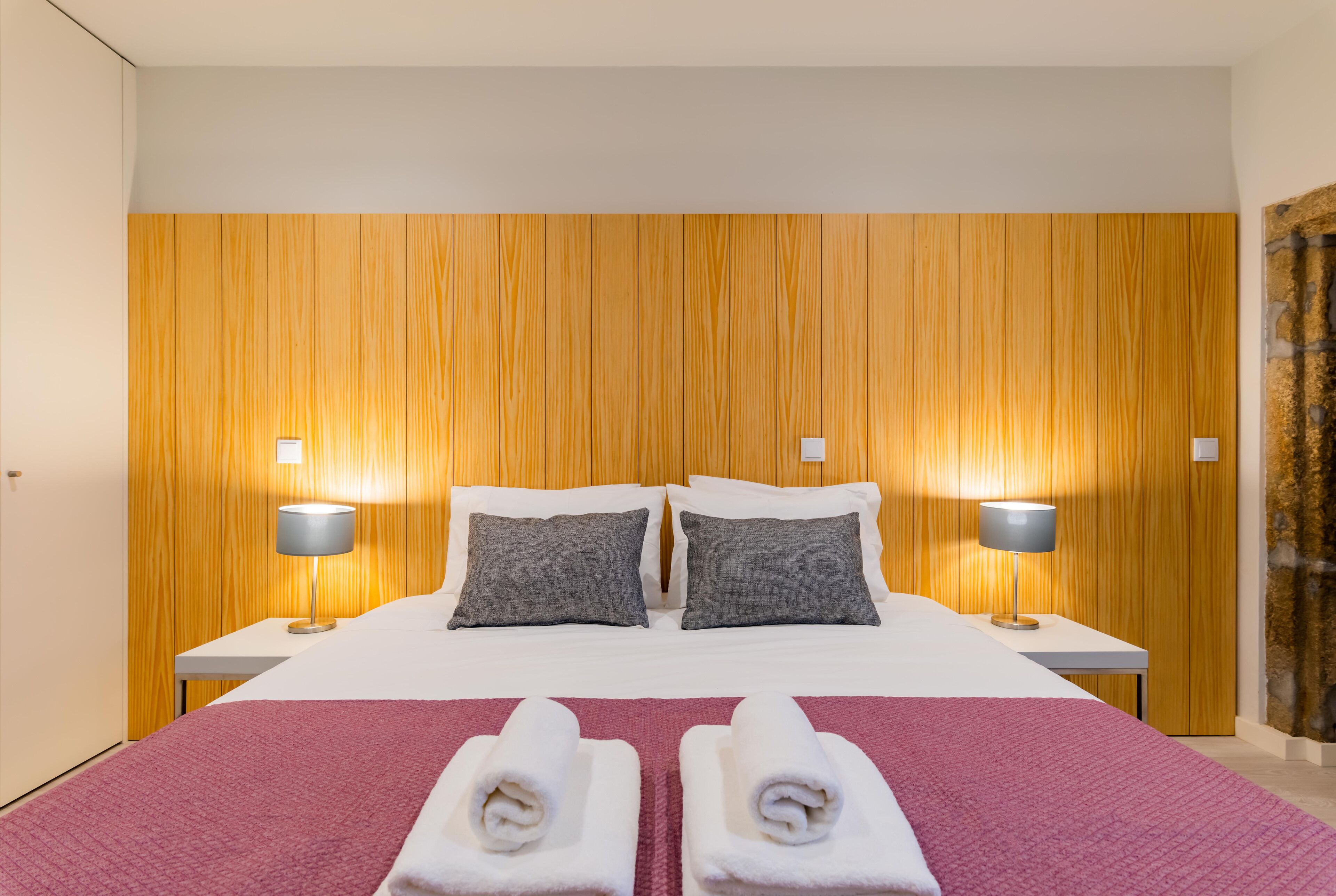 A hotel room with a wooden panel headboard, two bedside tables with lamps, a bed with a red blanket and two rolled towels, and a view of a stone wall.