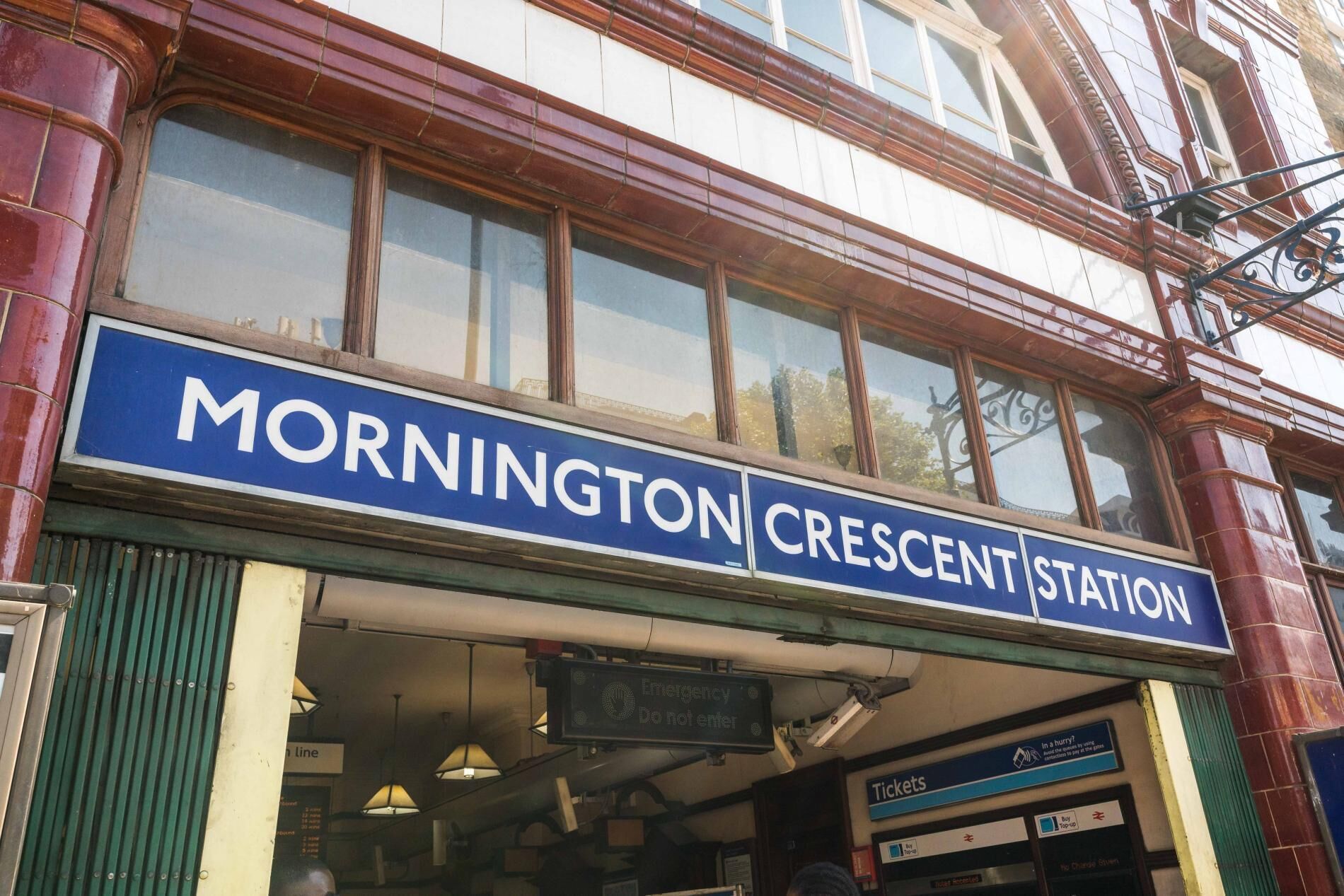 The exterior of Mornington Crescent Station with a blue signboard, emergency signage, and ticket office.