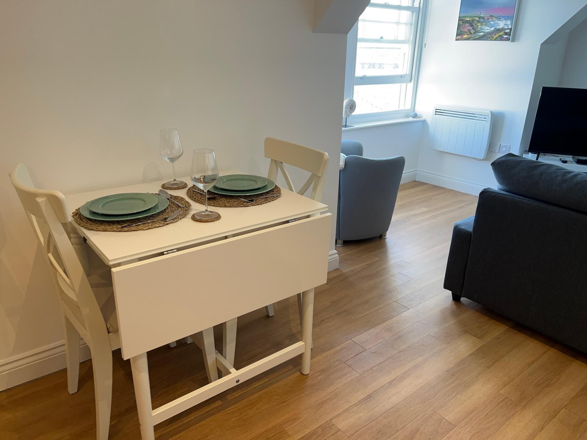 A dining area with a white table set for two, a grey sofa, and a TV in the background.