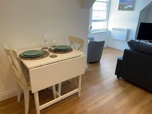 A dining area with a white table set for two, a grey sofa, and a TV in the background. - Apartment - Centre of Swanage Stunning Sea Views (Swanage)