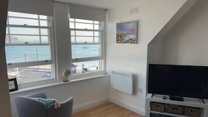 Ein Wohnzimmer mit großem Fenster mit Blick auf einen Hafen, einem Flachbildfernseher auf einem weißen Standfuß und einem grauen Sofa.