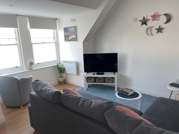 A modern living room with a grey sofa, a flat-screen TV on a white stand, a small side table with books, and a window with white blinds. - Apartment - Centre of Swanage Stunning Sea Views (Swanage)