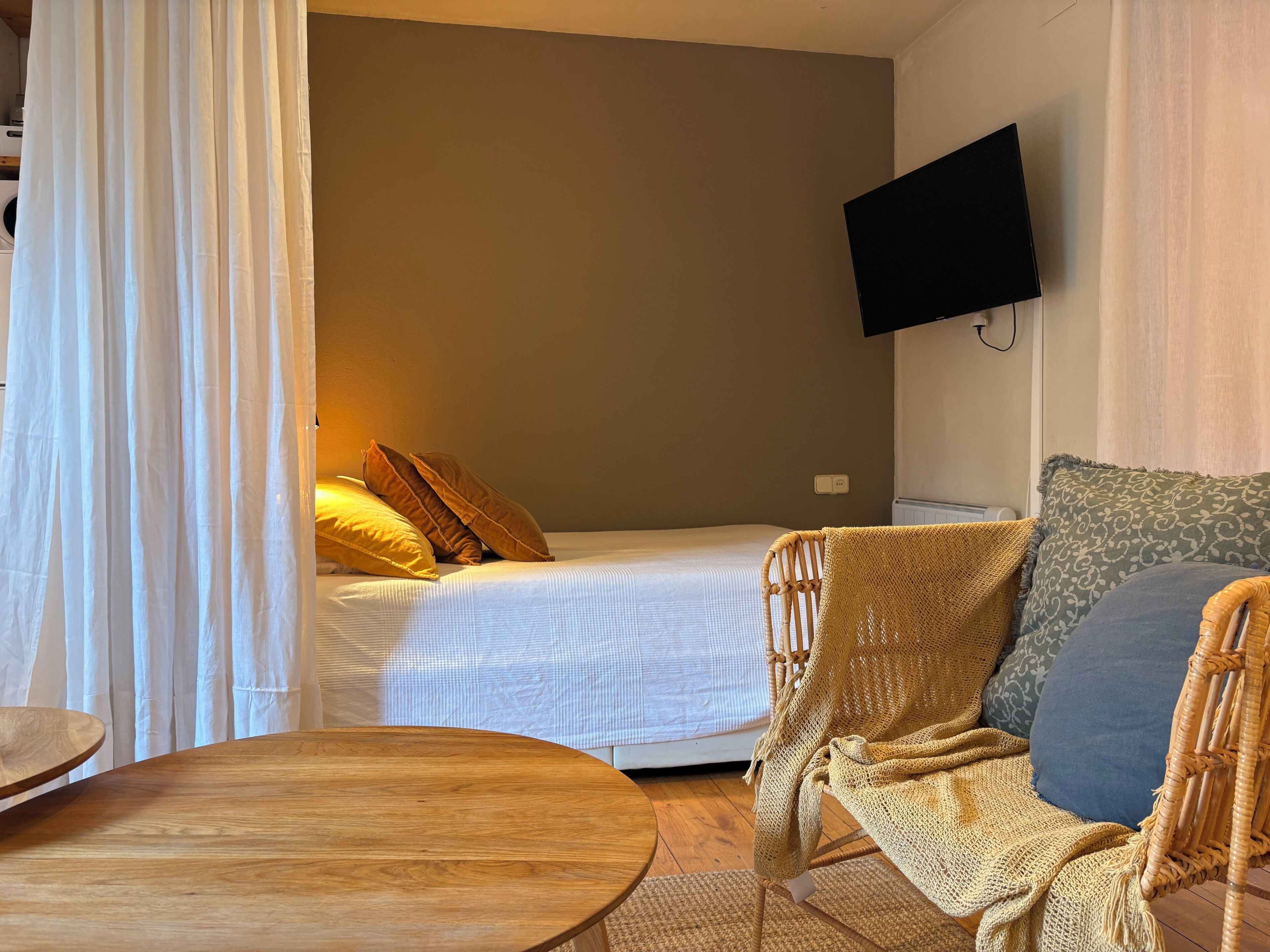A room with a bed, a television mounted on the wall, a wicker chair, and a round wooden table.