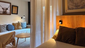 A cozy room with a bed, a small table, a chair, and a lamp. There are curtains and a framed picture on the wall.