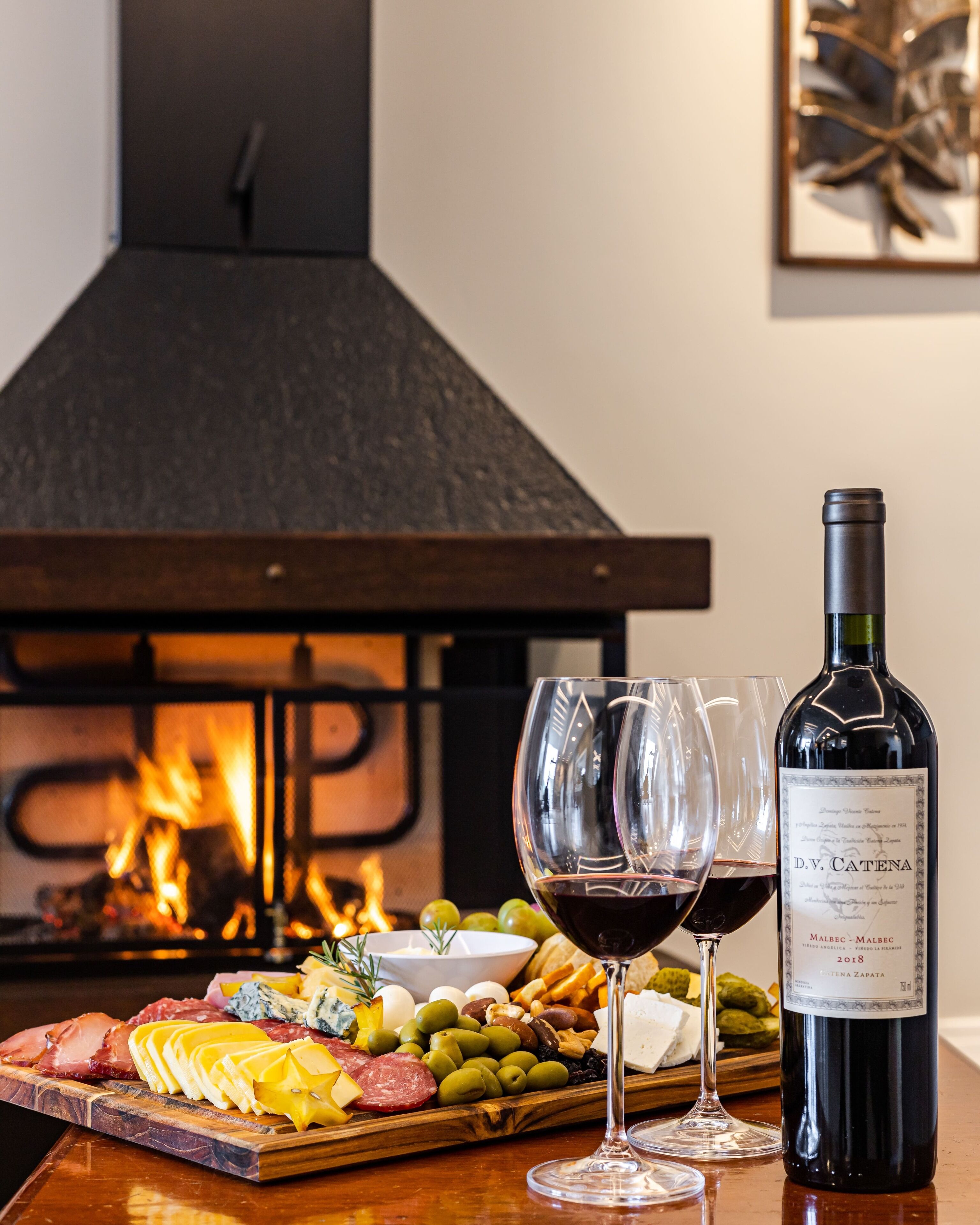 Una mesa de madera con una tabla de embutidos, dos copas de vino y una botella de vino.