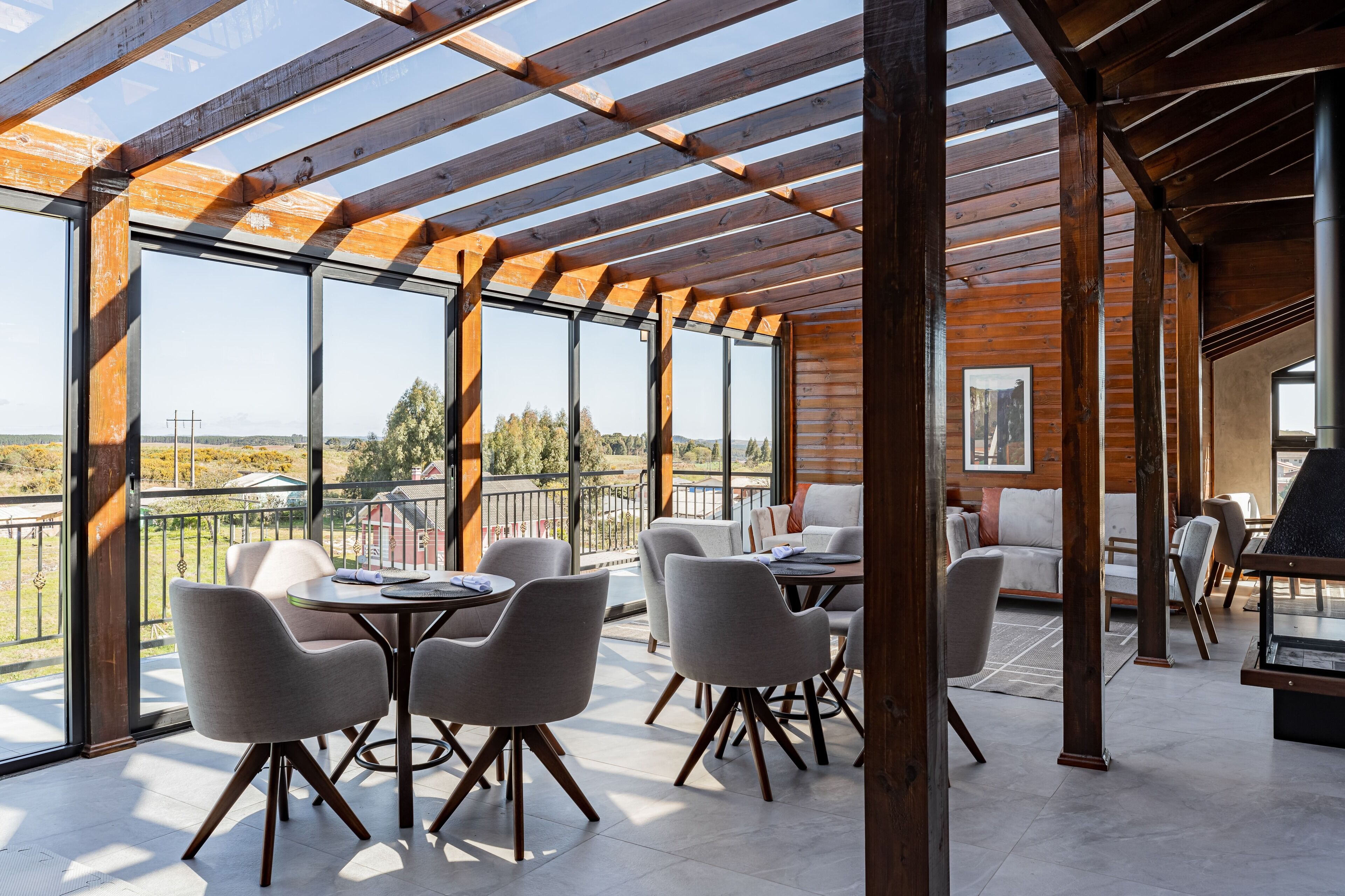 A spacious outdoor area with wooden beams and large windows, featuring a dining table and chairs, a sofa, and a fireplace.