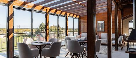 A spacious outdoor area with wooden beams and large windows, featuring a dining table and chairs, a sofa, and a fireplace.