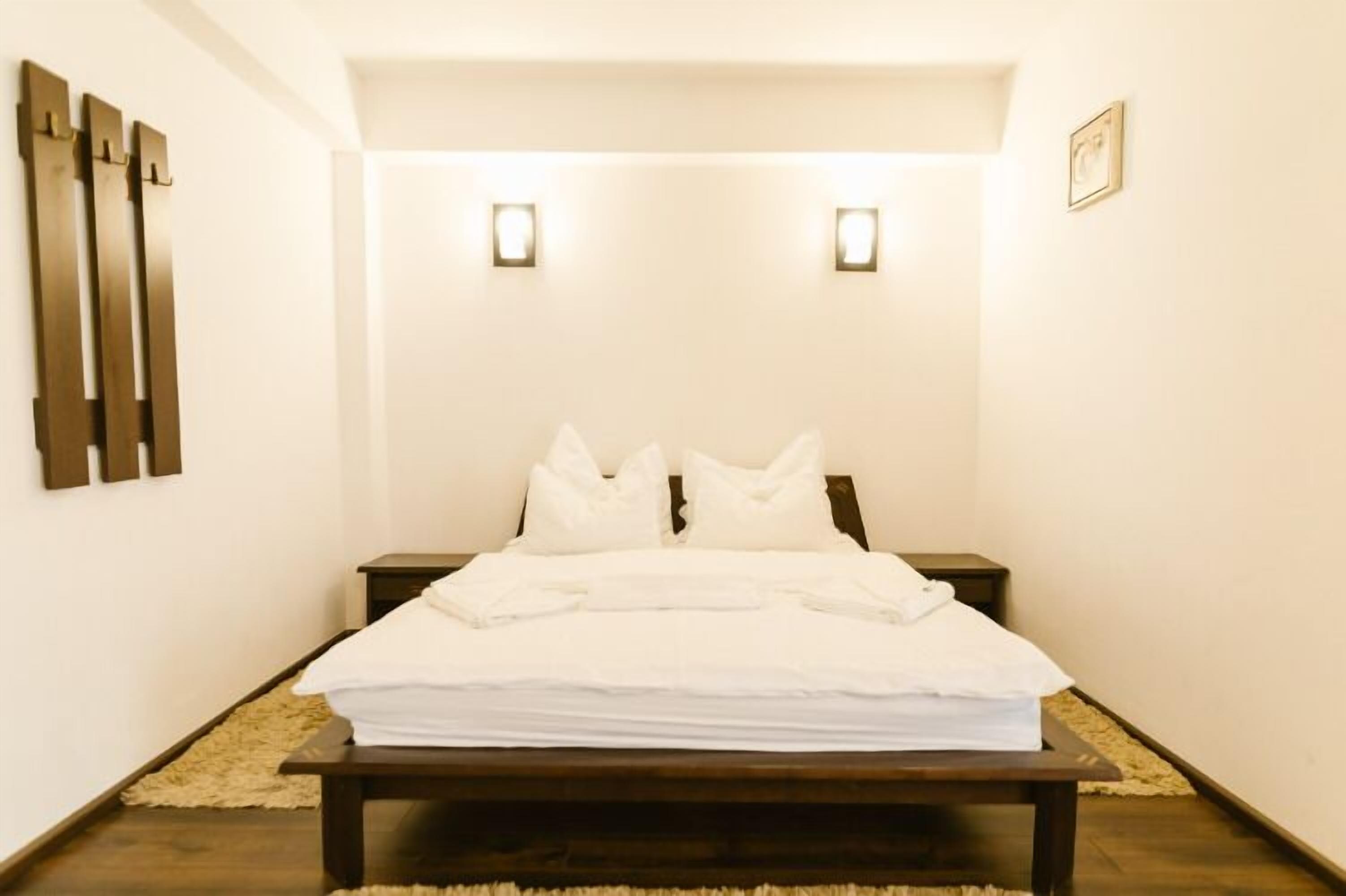A neatly made bed in a room with a simple, clean design. There are two bedside tables, one on each side of the bed. The walls are plain, and there are two small wall-mounted lights above the bed.