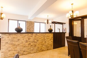 Un área de recepción con pared de piedra, dos jarrones grandes, una araña y una zona de estar con sillas.