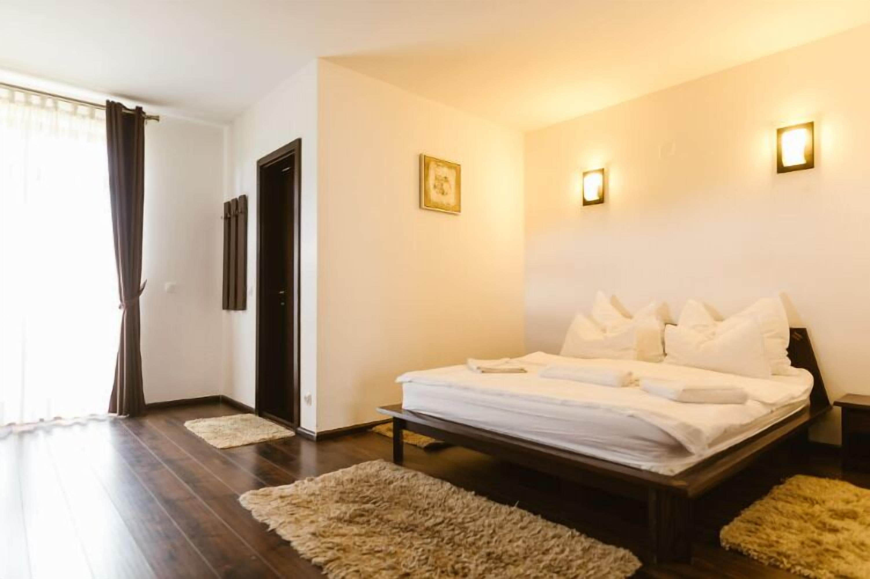 Un dormitorio con cama, dos luces fijadas a la pared, una puerta y un ventanal con cortinas.