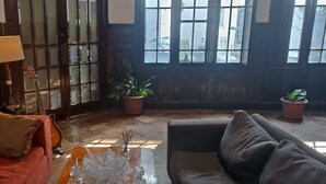 A cozy living room with a red sofa, a grey sofa, a wooden coffee table, and a guitar leaning against a wall. There are two potted plants and a window with a view of the outside.