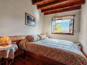 A bedroom with a wooden ceiling, a bed with floral bedding, a bedside table with a lamp, and a window with a view of trees and mountains.