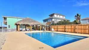 A swimming pool with a thatched-roof structure, a wooden fence, and houses in the background. - The Beach Buoy by Silver Sands Vacation Rentals (Port Aransas)